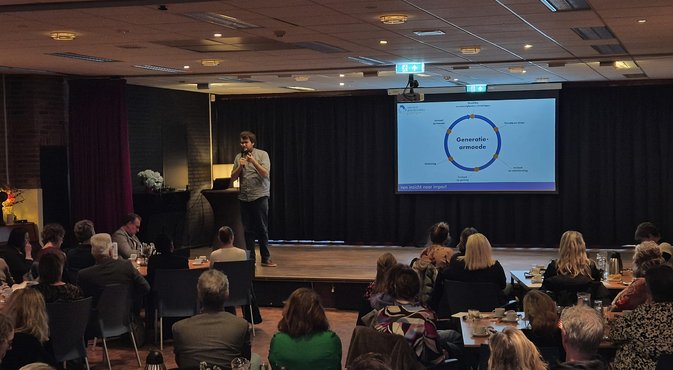 Foto: Zaal met publiek en spreker op podium